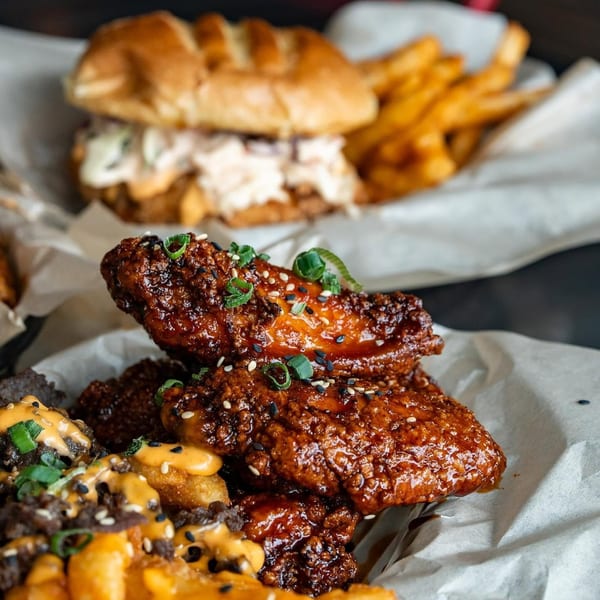 Crispy glazed chicken wings with sesame seeds and green onions, alongside loaded fries and a sandwich.