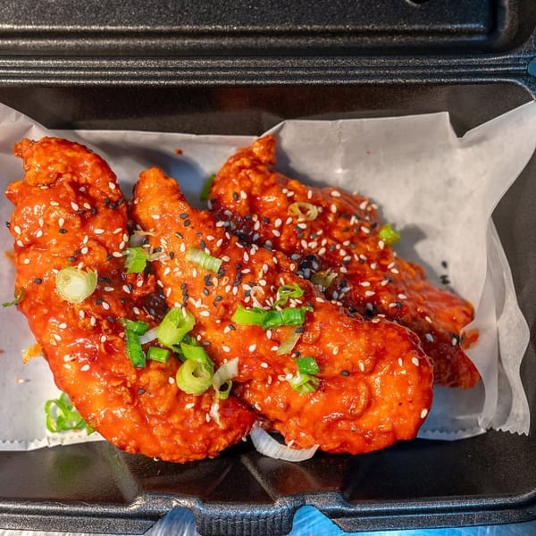 Crispy Korean fried chicken tenders in a black takeout container, garnished with sesame seeds and scallions.