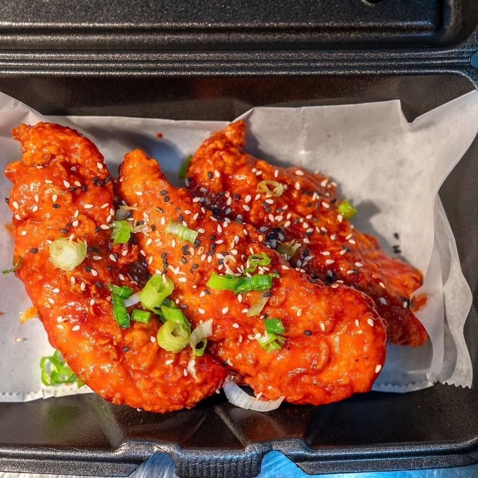 Crispy Korean fried chicken tenders in a black takeout container, garnished with sesame seeds and scallions.